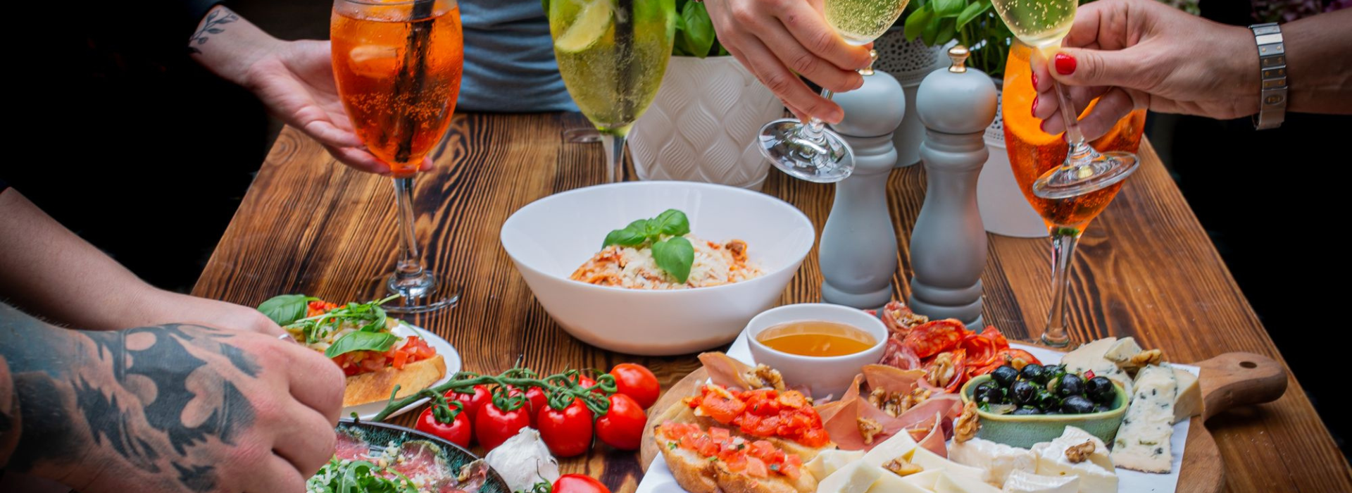 Wspólny stół w restauracji włoskiej w Dębkach, pełen antipasti, makaronu, bruschetty, serów, oliwek i kolorowych drinków – scena celebracji posiłku w letnim klimacie.